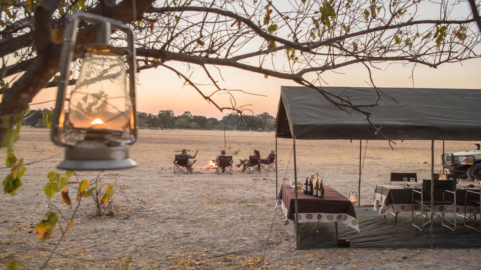 Camp dining table under a canvas awning, with guests around a fire at dusk